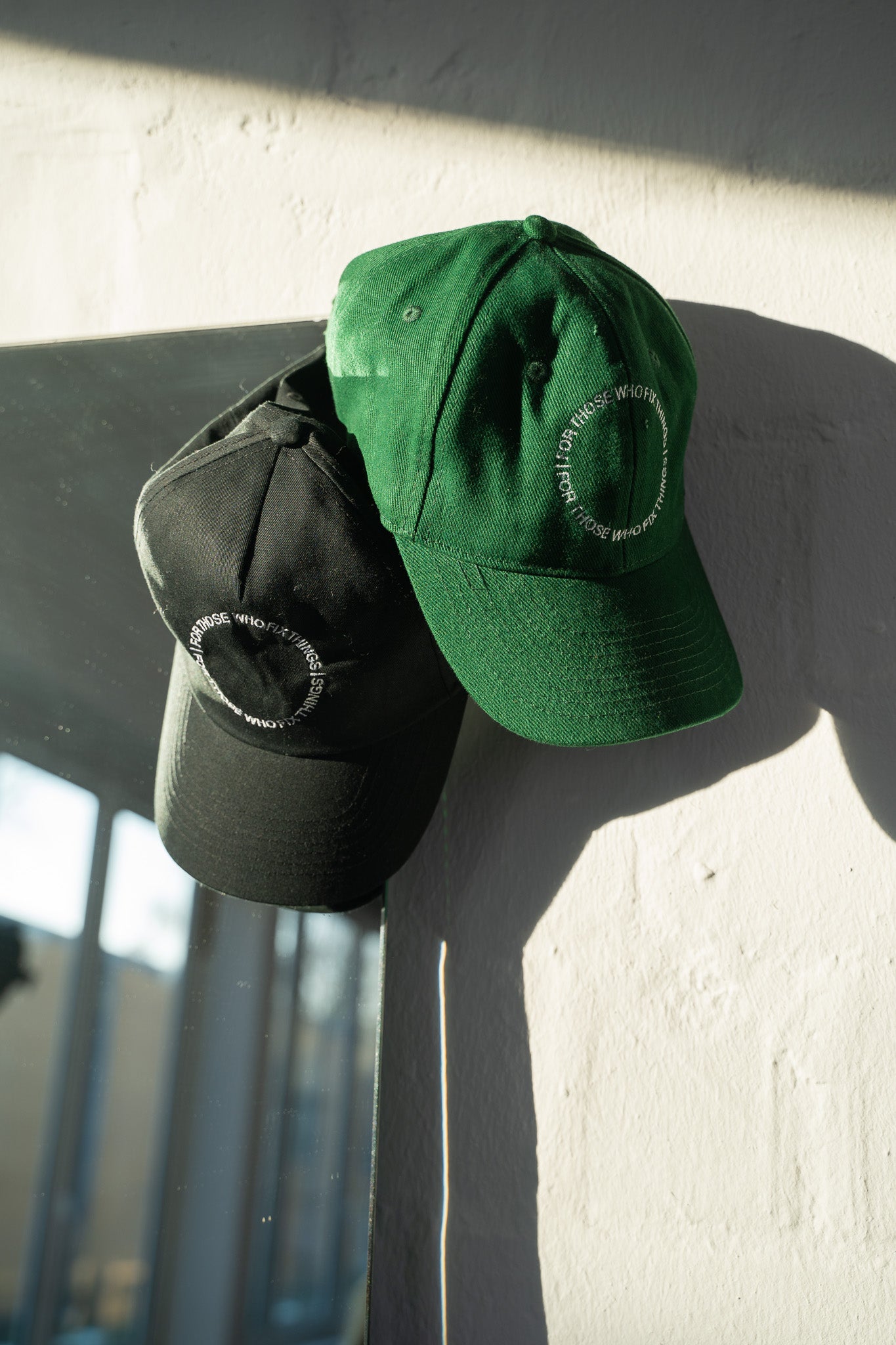 Two jan-n-june “CAP FOR THOSE WHO FIX THINGS BOTTLE GREEN GOTS” caps, crafted from organic cotton, sit on a black surface by a white wall. Each features an embroidered circular design with text. Sunlight creates shadows over the scene.