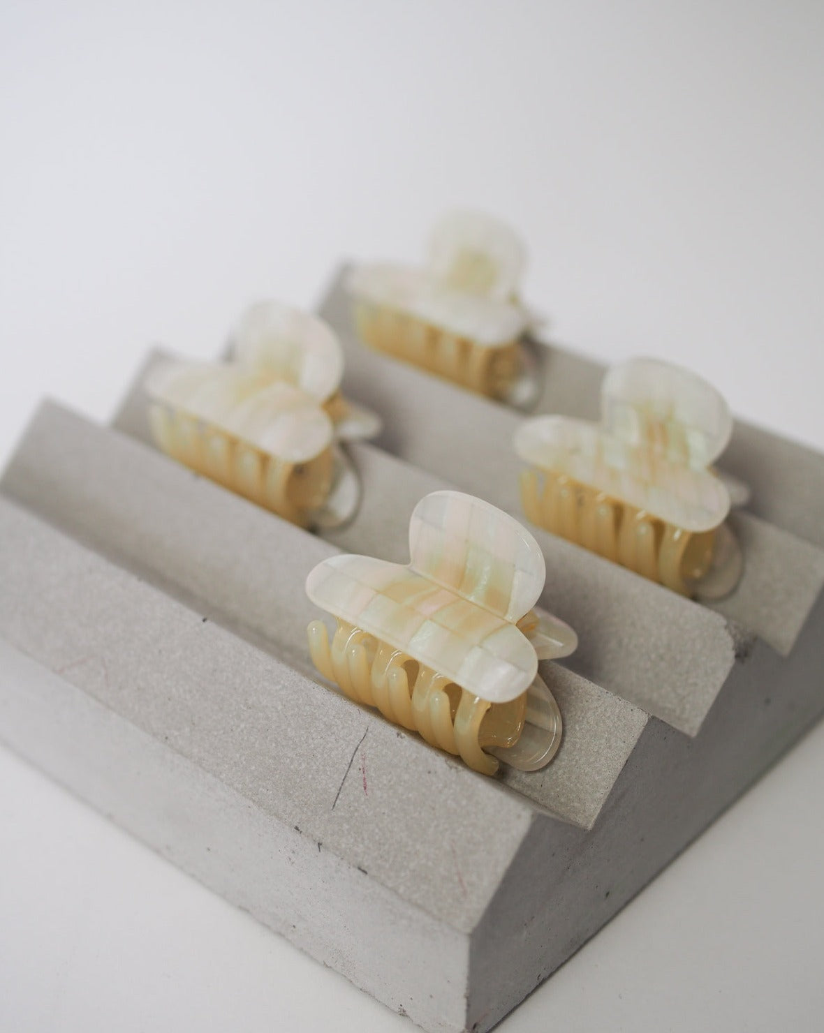 Four MINI HEIRLOOM CLIP OPALITE SHELL CHECKER hair claw clips by jan-n-june, in cream-toned iridescent acetate, are displayed on a gray stepped stand against a white background.