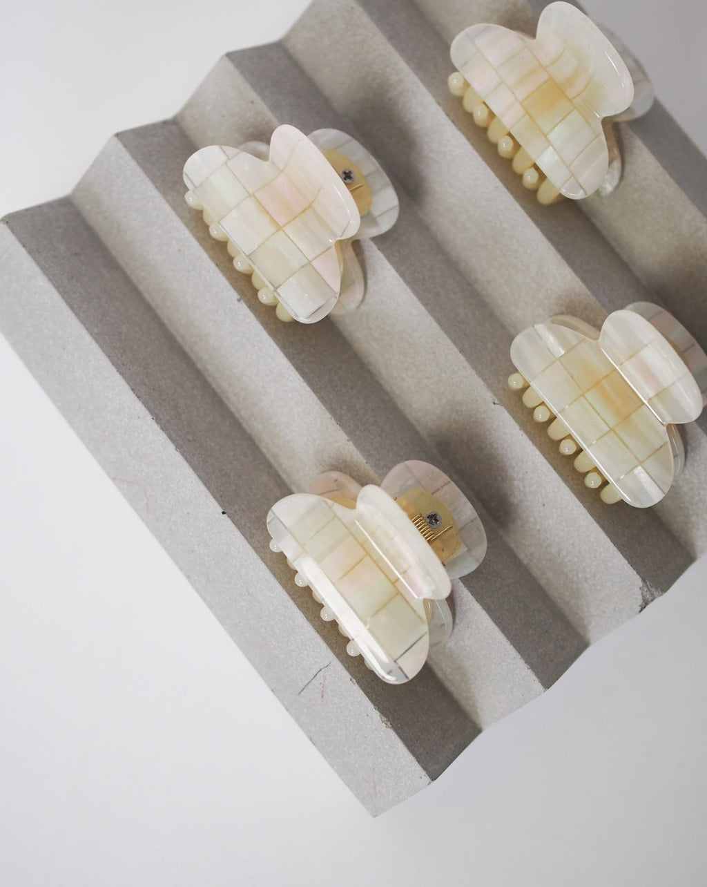 Four MINI HEIRLOOM CLIP OPALITE SHELL CHECKER hair claws by jan-n-june are neatly lined up on a textured, wavy gray stand, viewed from above against a plain light background.
