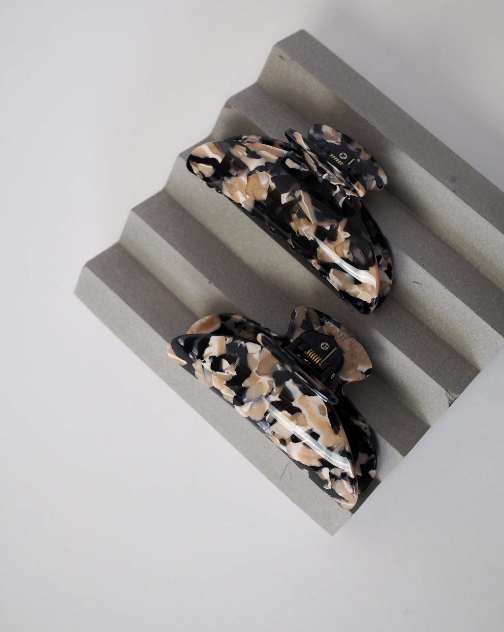 Two MIDI HEIRLOOM CLAW ABALONE clips by jan-n-june, crafted from Italian acetate in black, beige, and cream marbled tones, are displayed on a gray wavy-textured surface against a plain white background.