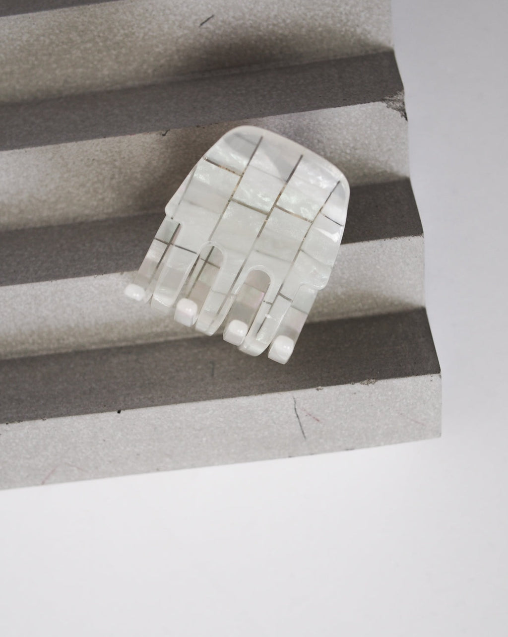 The MINI CLAW OPALITE SHELL CHECKER by jan-n-june is a small, abstract, hand-shaped accessory made from shiny white segmented material, displayed on a gray ridged surface with a plain background.