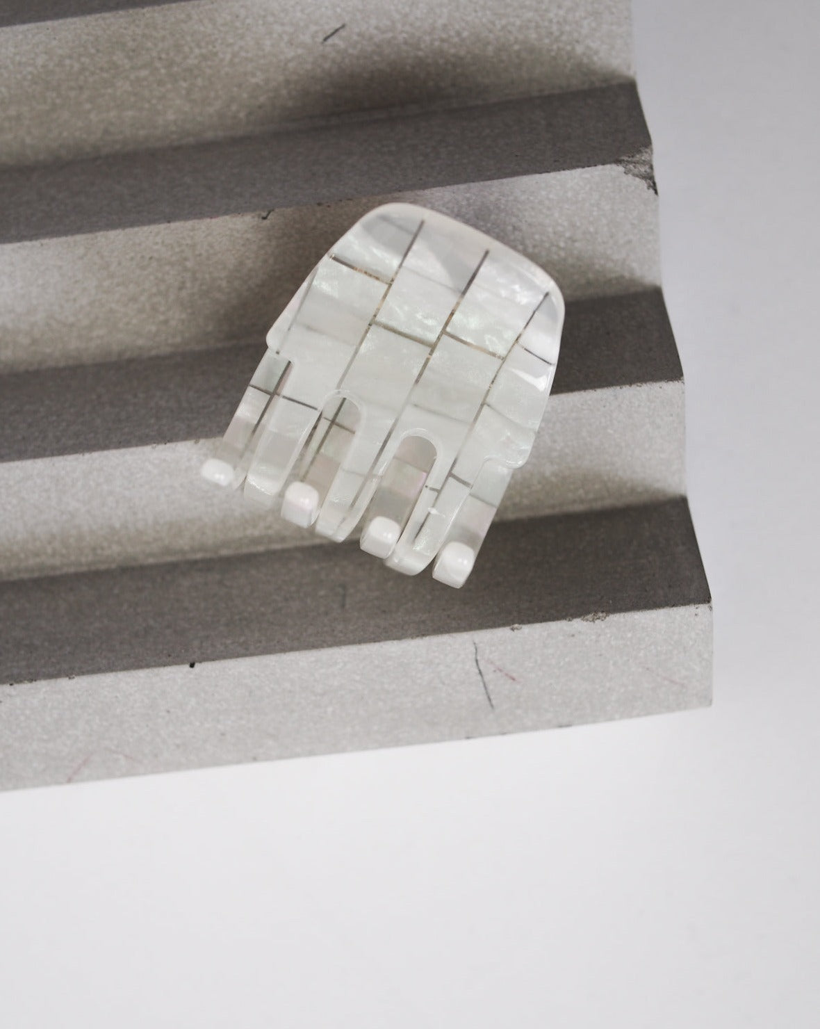The MINI CLAW OPALITE SHELL CHECKER by jan-n-june is a small, abstract, hand-shaped accessory made from shiny white segmented material, displayed on a gray ridged surface with a plain background.