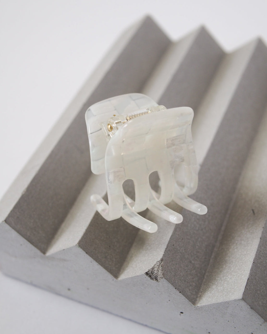 The MINI CLAW OPALITE SHELL CHECKER by jan-n-june, a translucent, handcrafted rectangular hair claw clip with a marble-like checker pattern, rests on a gray ridged geometric surface.