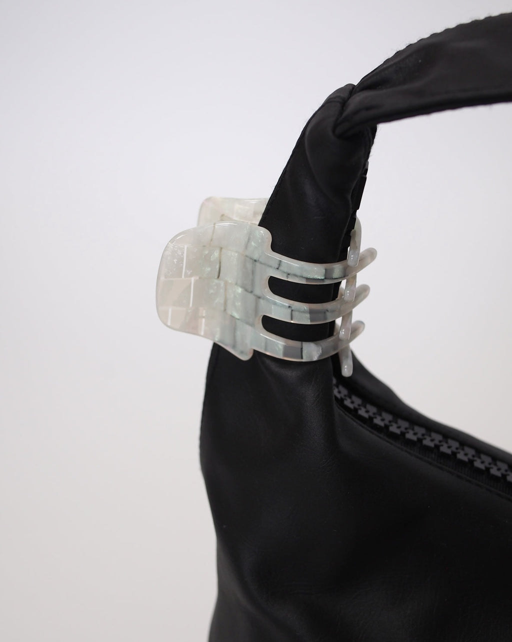 A black bag with the jan-n-june MINI CLAW OPALITE SHELL CHECKER, a marbled translucent white hair claw handcrafted in Italy, clipped to its strap. Set against a plain white background.