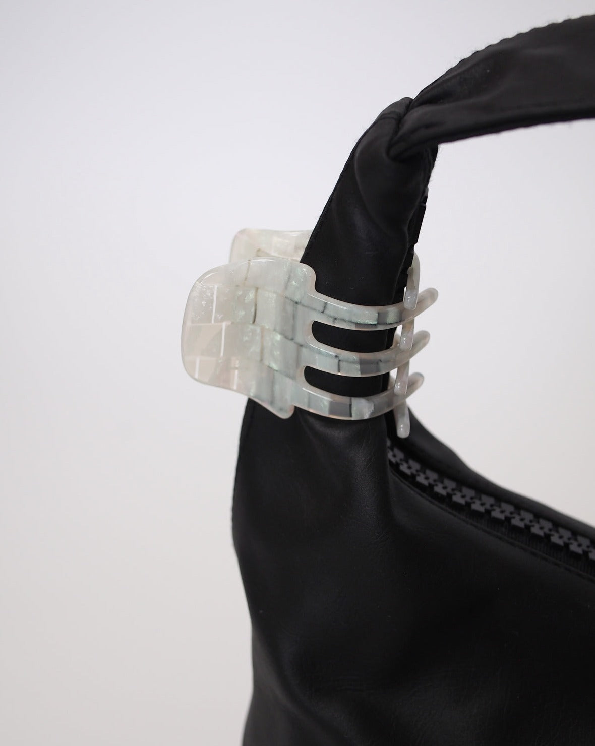 A black bag with the jan-n-june MINI CLAW OPALITE SHELL CHECKER, a marbled translucent white hair claw handcrafted in Italy, clipped to its strap. Set against a plain white background.