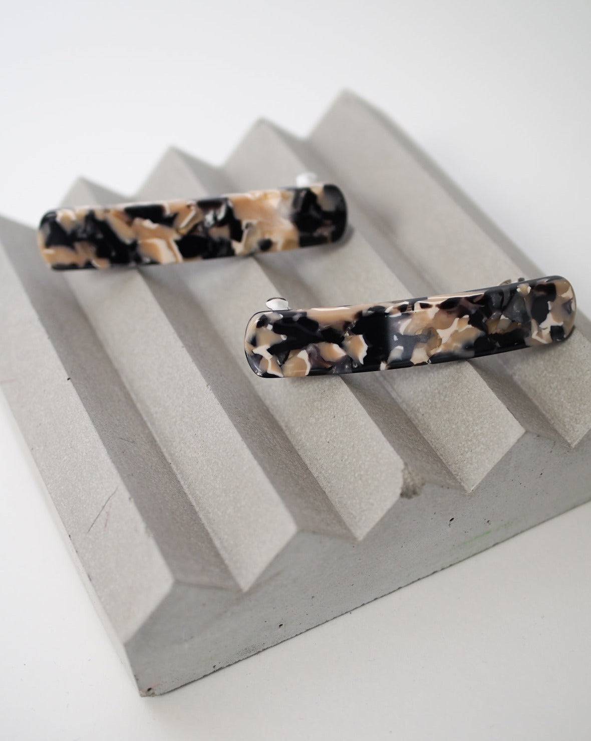 Two HEIRLOOM BARRETTE ABALONE hair clips by jan-n-june, made from premium Italian acetate in a marbled black, beige, and cream pattern, are displayed on a gray zigzag stand.