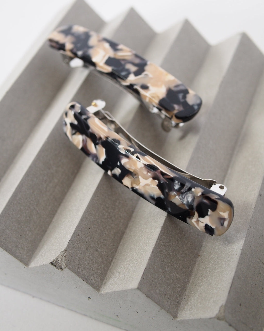 Two rectangular HEIRLOOM BARRETTE ABALONE hair clips by jan-n-june, featuring a marbled black, beige, and cream Italian acetate design, are displayed on a textured light gray ridged surface.