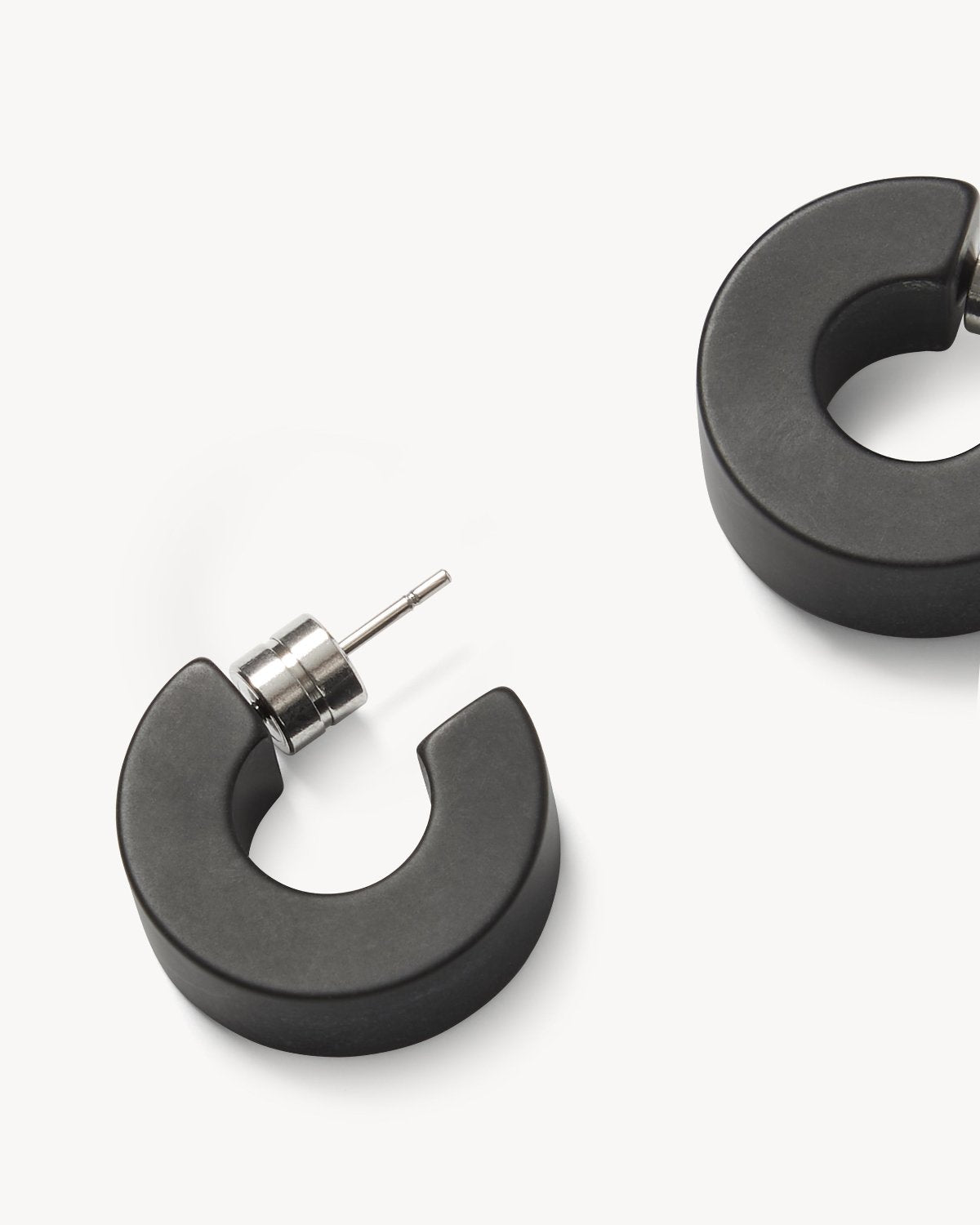 Close-up of two chunky, black matte jan-n-june MUSE HOOPS with silver posts on a white background. One sustainable earring is fully visible while the other is partially shown—perfect for everyday wear.