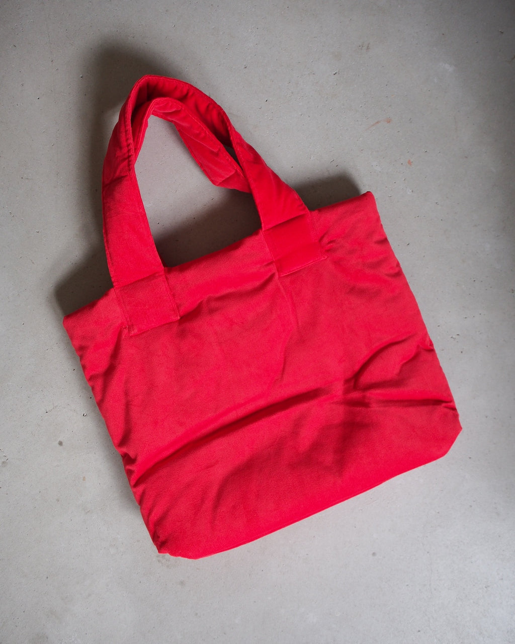 The jan-n-june PADDED SHOPPER SMOL RED, a bright red tote bag made from organic cotton, lies flat on a light gray textured surface.