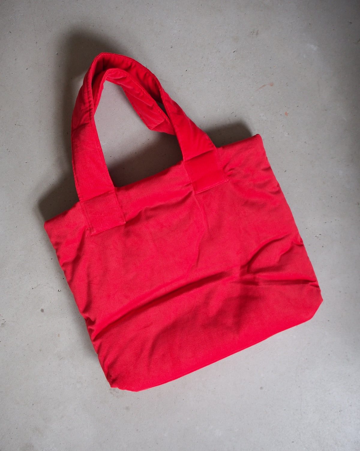 The jan-n-june PADDED SHOPPER SMOL RED, a bright red tote bag made from organic cotton, lies flat on a light gray textured surface.