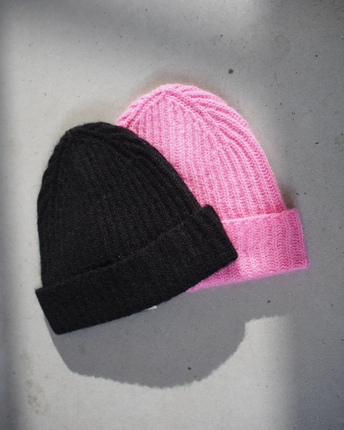 The jan-n-june WOOLY KNIT BEANIE BJON BLACK and a pink version, both crafted from a mohair and wool blend, rest on a light gray surface. Each ribbed, folded-brim beanie features a cozy yet stylish texture, with the pink beanie slightly overlapping the black.