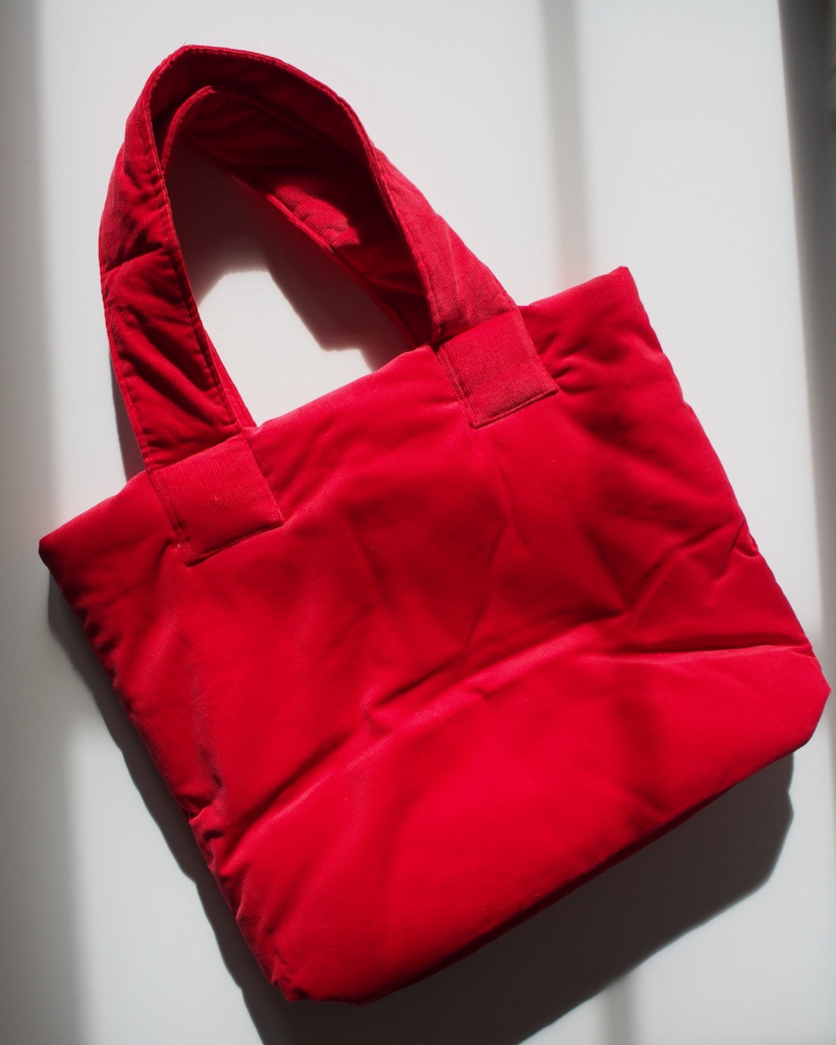The jan-n-june PADDED SHOPPER SMOL RED, an organic cotton padded tote with two handles, is displayed on a white surface with soft overhead shadows.