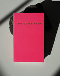 The KALENDER EIN GUTER PLAN FLAMINGO UNDATED by jan-n-june, with its bright pink hardcover and gold title, rests partly on a dark rock amid dramatic shadows—ideal for mindfulness and self-love.
