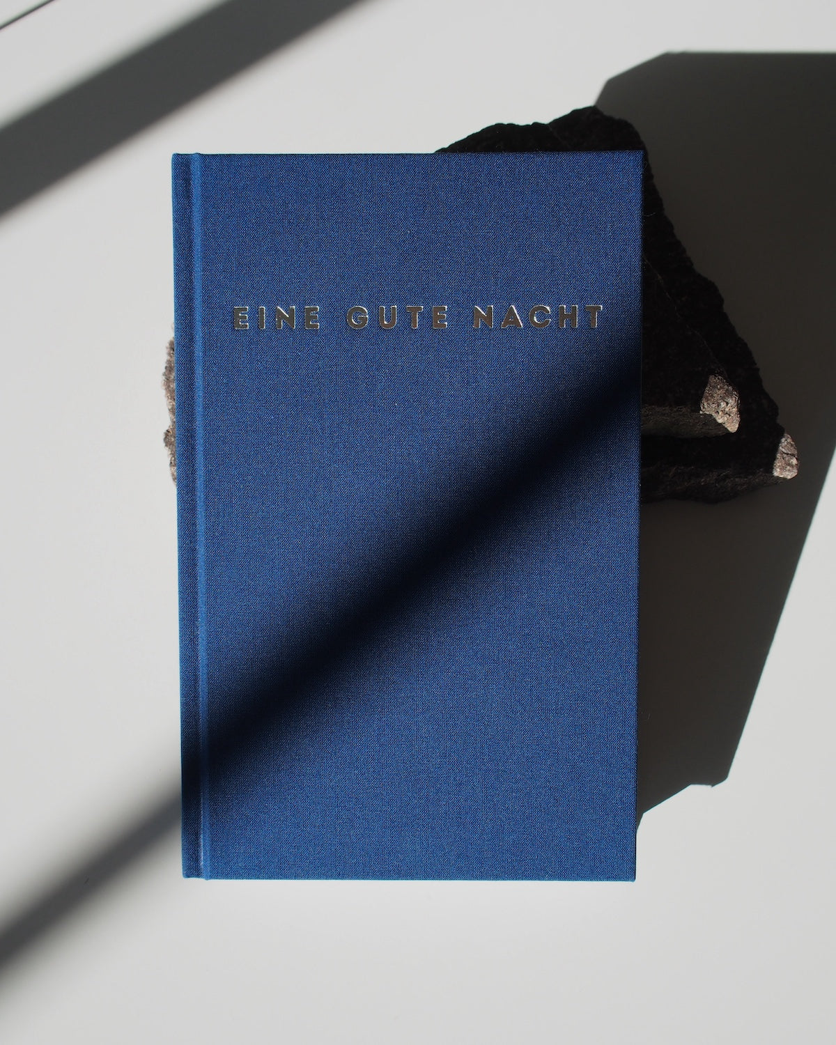 The jan-n-june TAGEBUCH EINE GUTE NACHT is a blue sleep diary with gold embossed text on the cover, resting on a dark textured surface and inviting mindful reflection as gentle shadows fall across the scene.