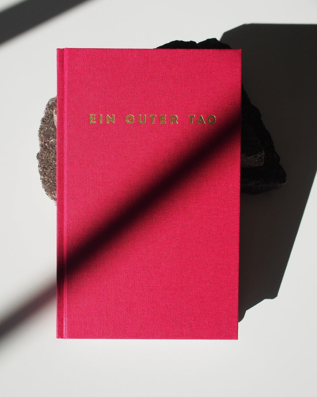 The TAGEBUCH EIN GUTER TAG RUBIN by jan-n-june, with gold lettering on bright pink, rests on a dark rock atop a white surface, its cover partly shaded—inviting mindfulness and self-love.