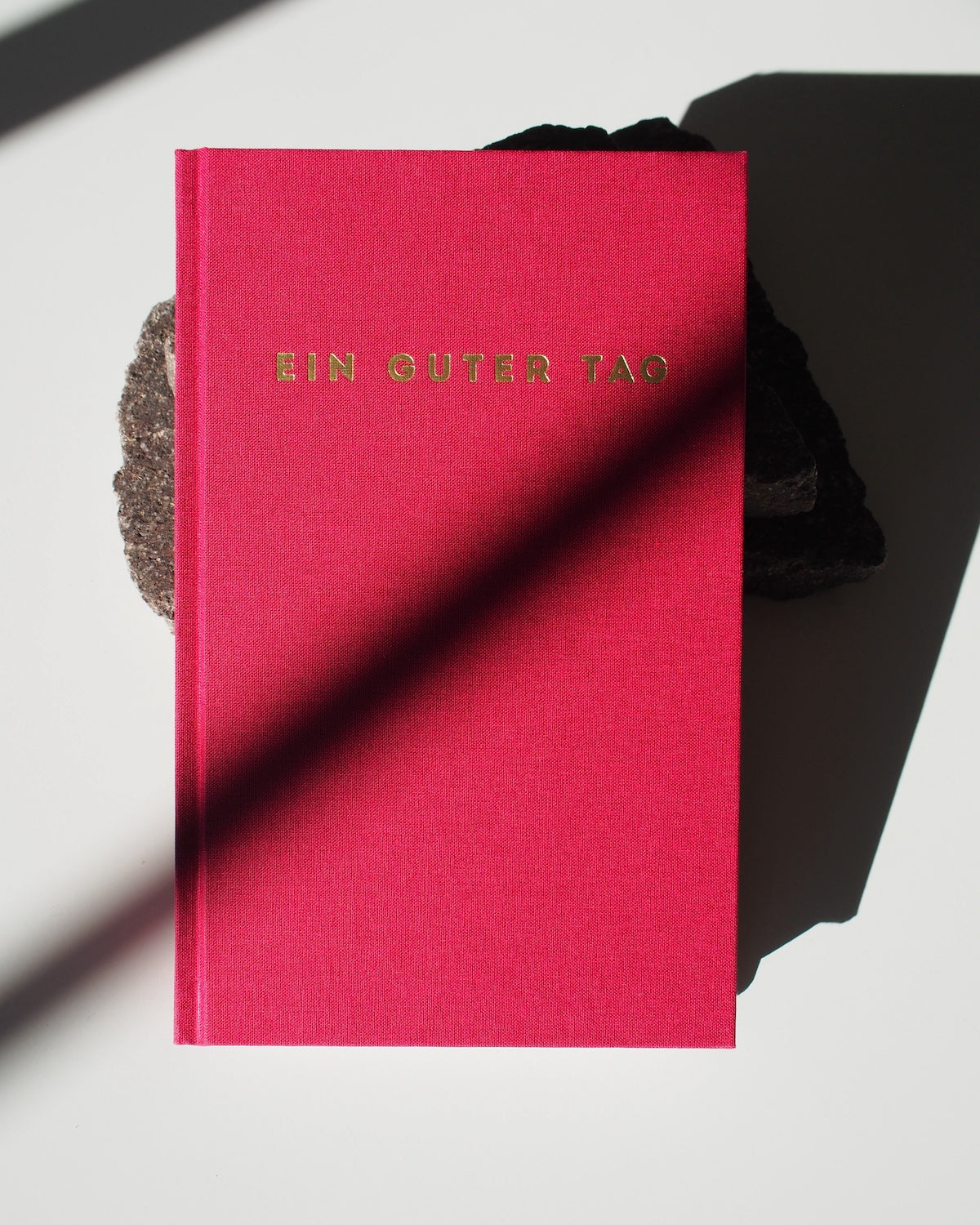 The TAGEBUCH EIN GUTER TAG RUBIN by jan-n-june, with gold lettering on bright pink, rests on a dark rock atop a white surface, its cover partly shaded—inviting mindfulness and self-love.