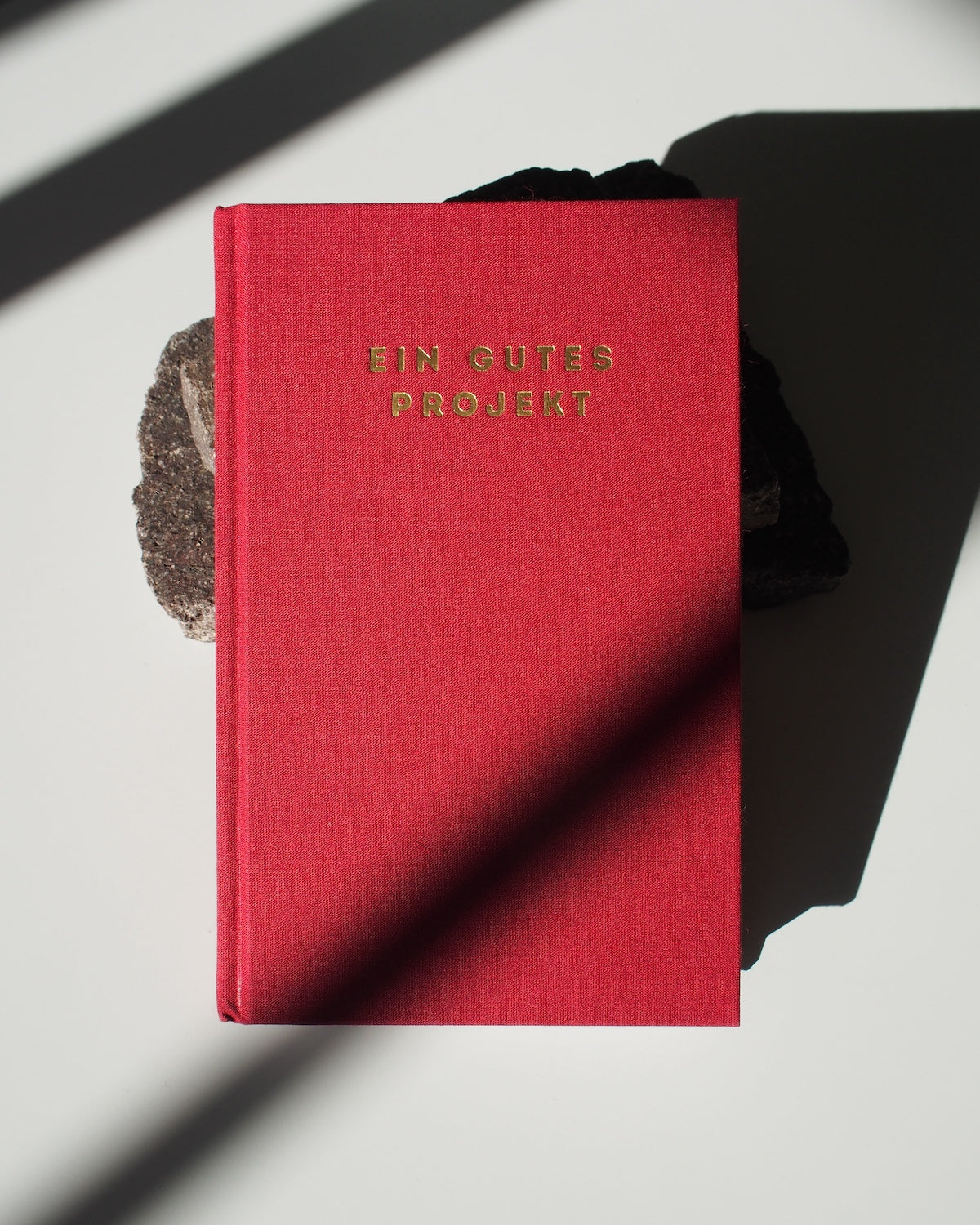 The jan-n-june PLANER EIN GUTES PROJEKT RUBIN, a red hardcover mindful success journal in gold lettering, rests partly on a dark rock atop a white surface, with diagonal shadows crossing the image.