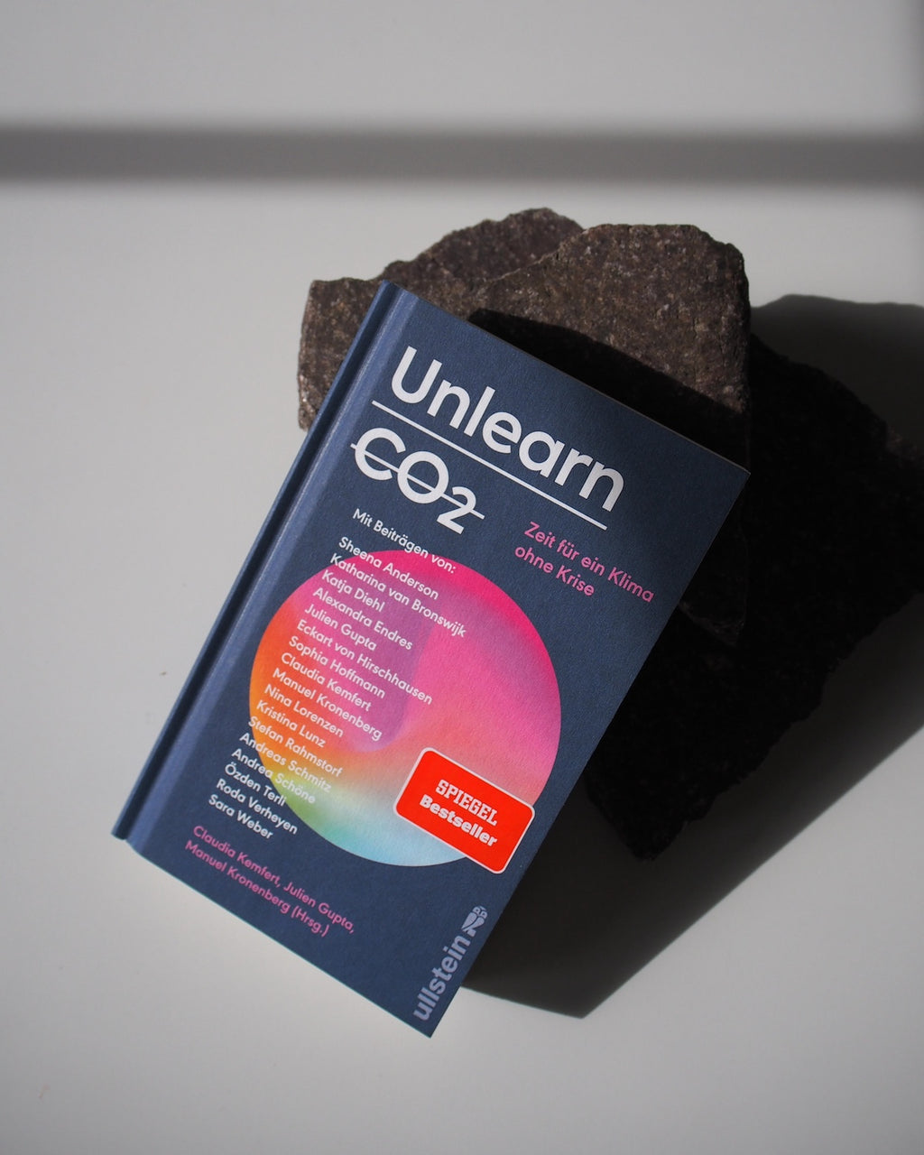 The book UNLEARN CO2 by jan-n-june, featuring a colorful circular cover design and a red “SPIEGEL Bestseller” sticker, sits on a rough dark stone slab in bright sunlight, highlighting climate-friendly future themes and multiple authors.