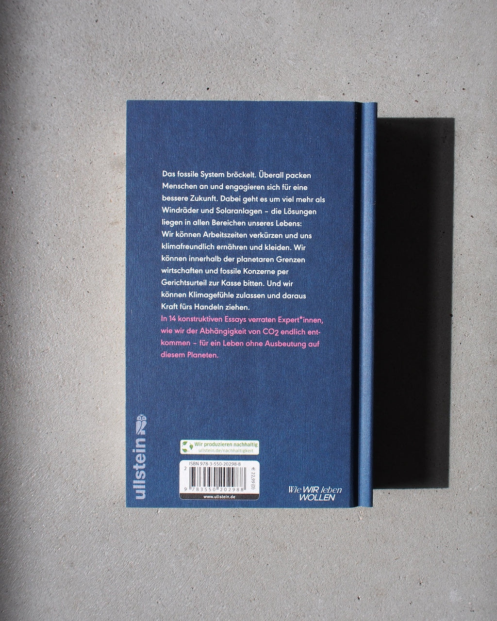 The back cover of the blue UNLEARN CO2 book by jan-n-june discusses fossil fuel systems, climate change, and urgent action. Sunlight highlights its call for a climate-friendly future as it rests on a gray surface.