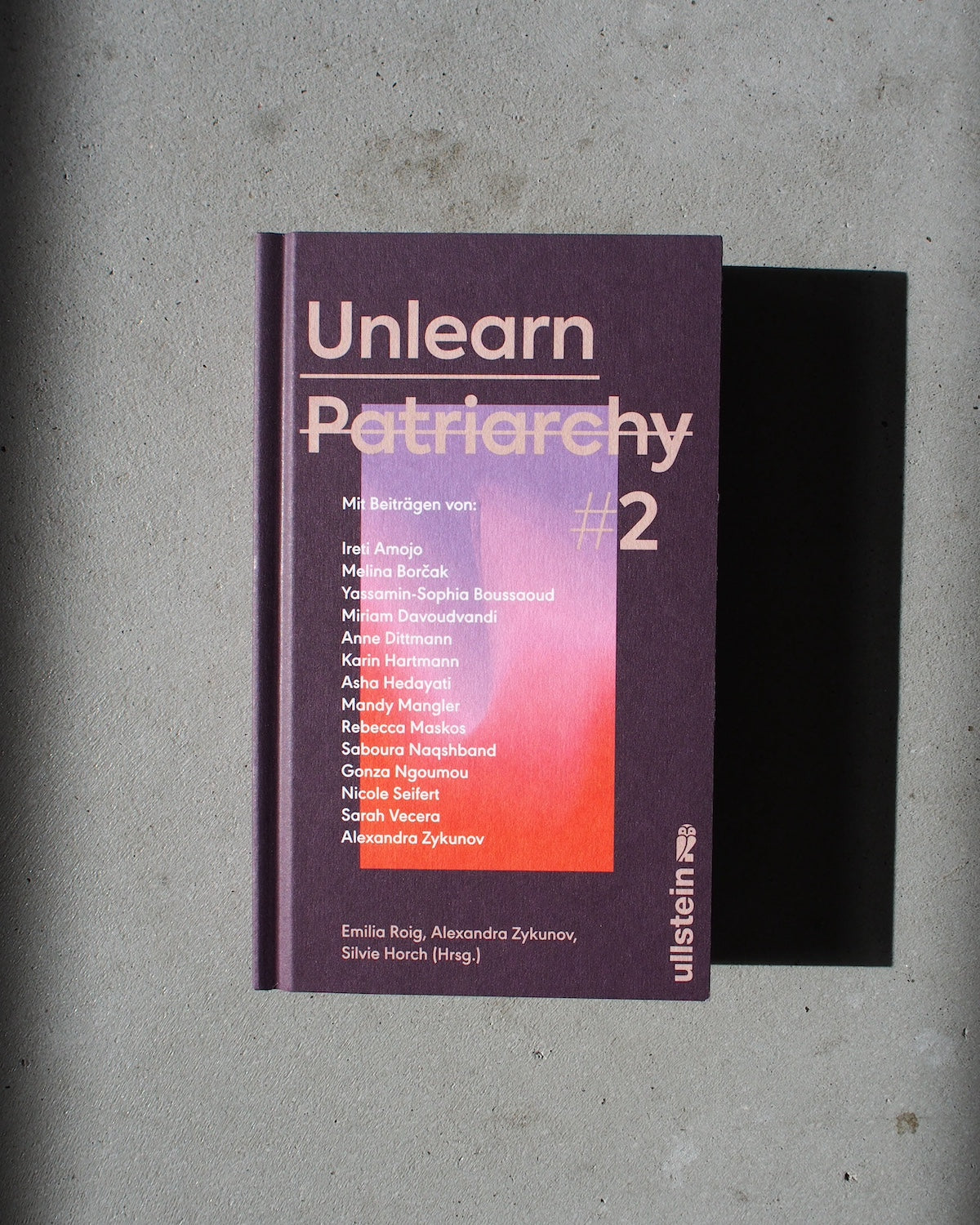 A purple hardcover book titled UNLEARN PATRIARCHY 2 by jan-n-june rests on a gray surface, featuring white and pink contributor names on the cover. The publisher is Ullstein, shown at the bottom right.