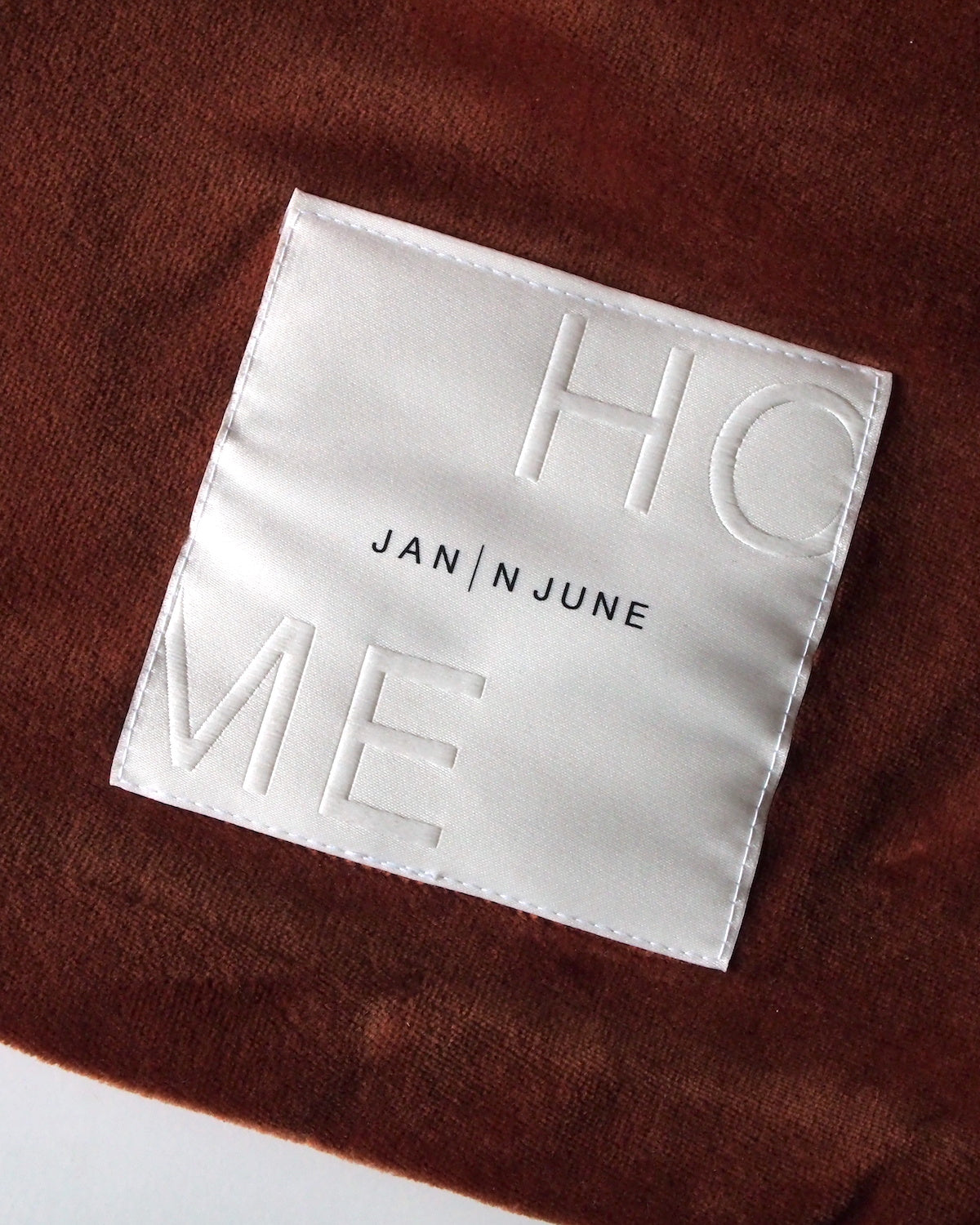 A white organic cotton patch with embossed HOME and jan-n-june text is sewn onto the HOT WATER BOTTLE COVER NICKI BRICK, adding a sustainable accent to the brown textured design.