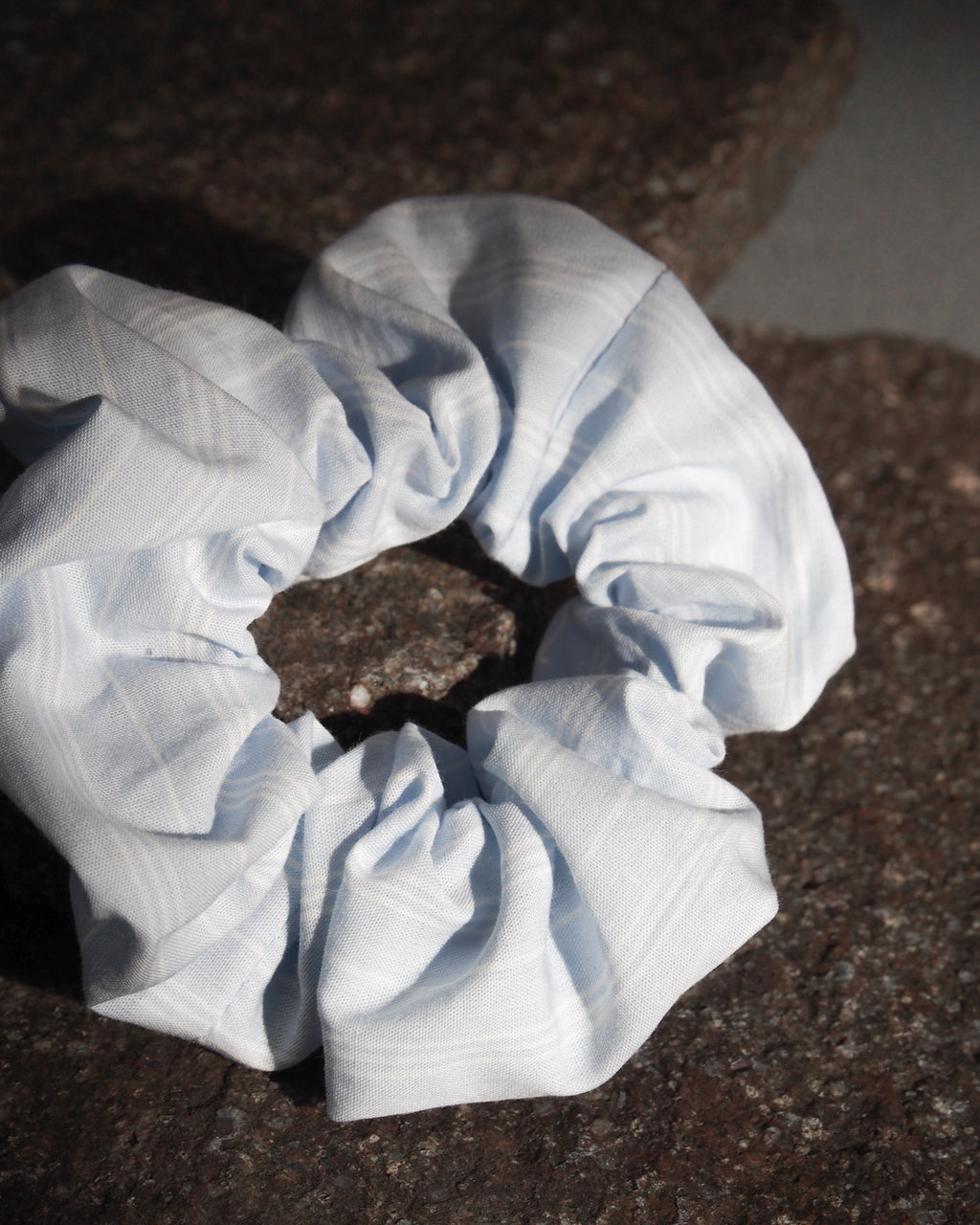 The SCRUNCHIE BLUE WHITE STRIPED by jan-n-june, made from offcuts, features blue and white stripes and rests on a rugged dark stone surface, softly illuminated by natural light.