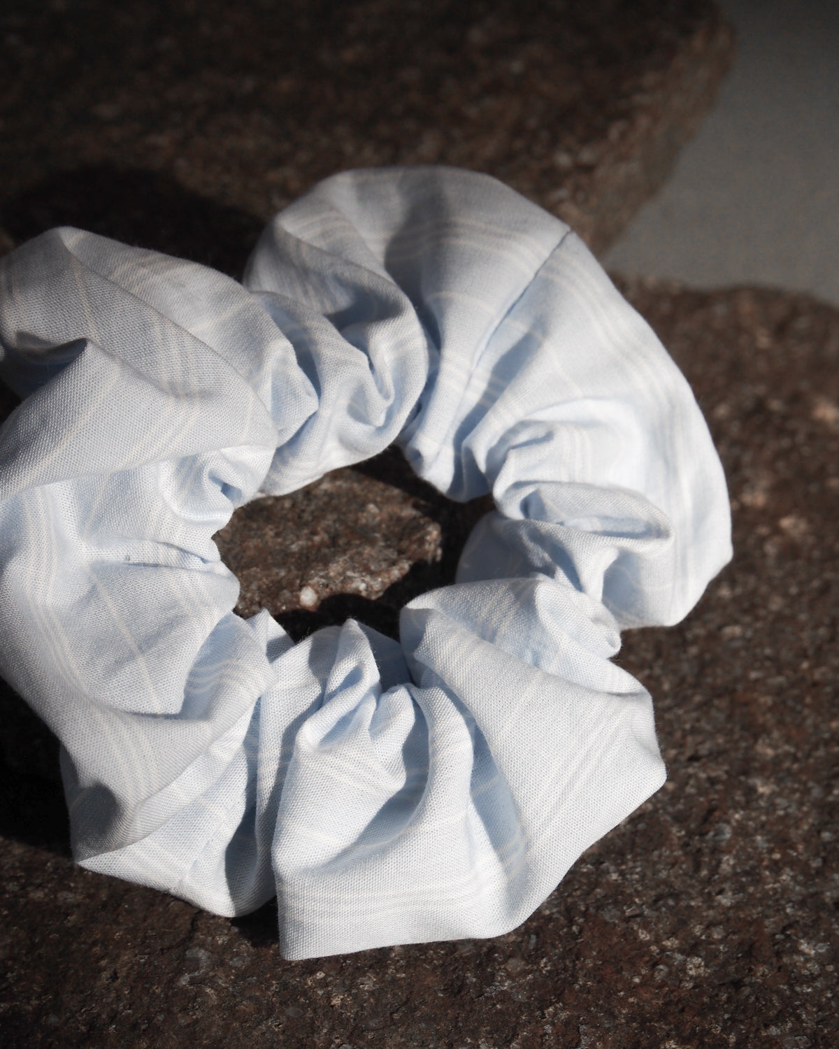 The SCRUNCHIE BLUE WHITE STRIPED by jan-n-june, made from offcuts, features blue and white stripes and rests on a rugged dark stone surface, softly illuminated by natural light.