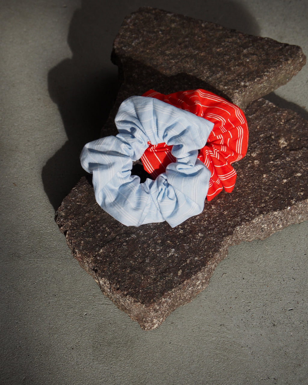 Two jan-n-june SCRUNCHIE BLUE WHITE STRIPED accessories, crafted from offcuts—one light blue with thin stripes and one red with white stripes—rest on a jagged stone surface against a gray background.