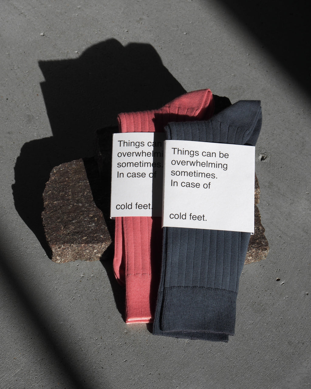Two pairs of jan-n-june SOCKS BERRY, one pink and one dark gray, made from organic cotton, rest on rough stone. Each pair features a label: Things can be overwhelming sometimes. In case of cold feet.
