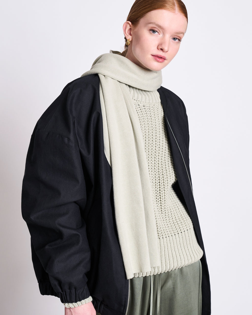 A person with fair skin and red hair wears the jan-n-june KNIT SCARF STATEN VINTAGE LACE GOTS, gold hoops, a cream knit sweater, oversized black jacket, and green satin pants against a plain white background.