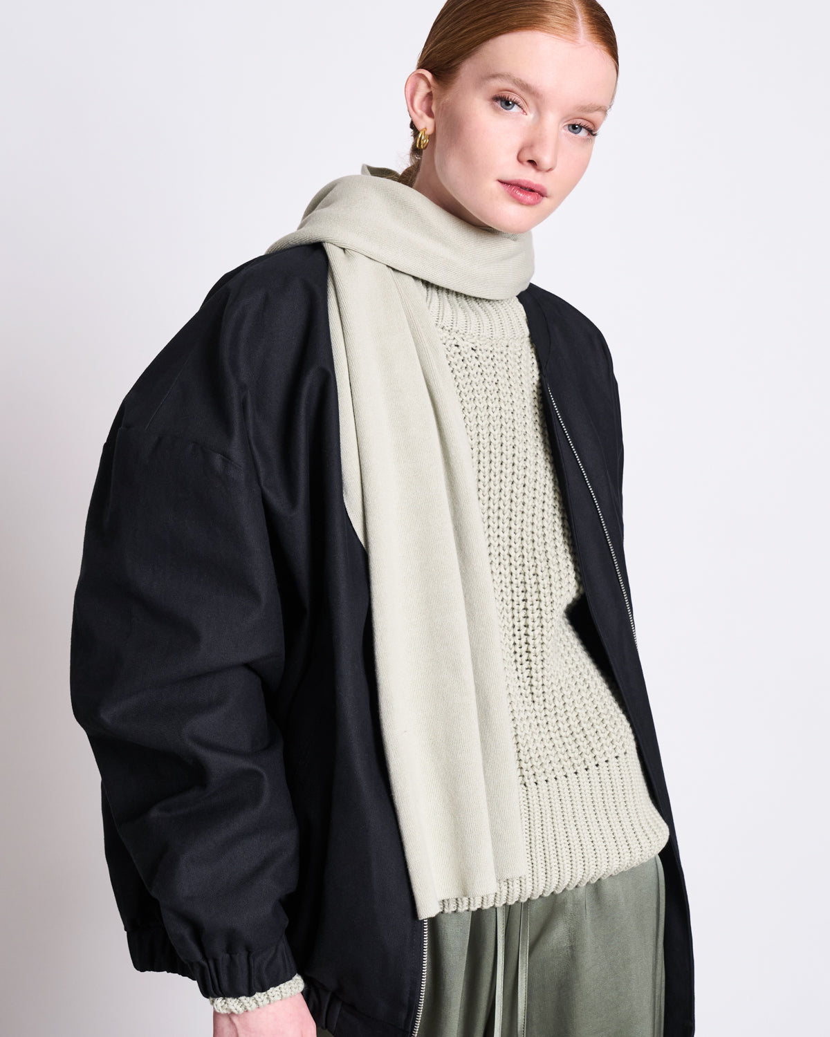 A person with fair skin and red hair wears the jan-n-june KNIT SCARF STATEN VINTAGE LACE GOTS, gold hoops, a cream knit sweater, oversized black jacket, and green satin pants against a plain white background.