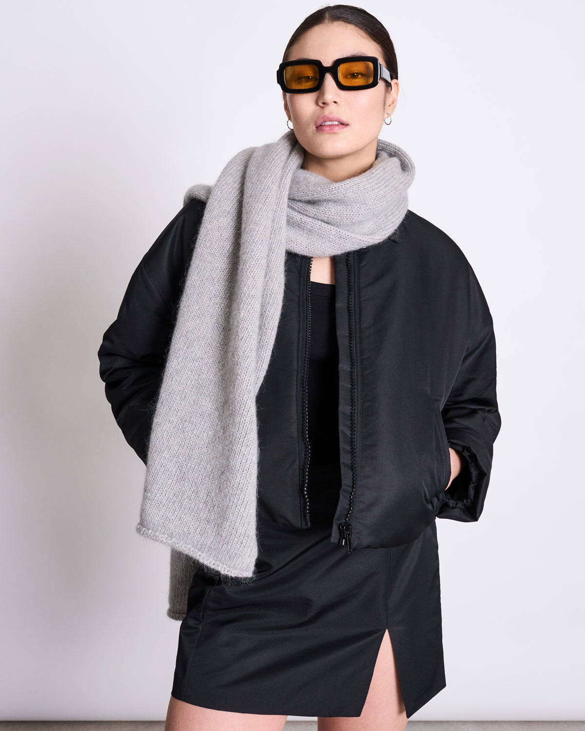 A woman in a black jacket, mini dress with front slit, and amber sunglasses stands against a light background, wrapped in the cozy jan-n-june WOOLY KNIT SCARF AVAK LIGHT GREY crafted from a soft wool and mohair blend.