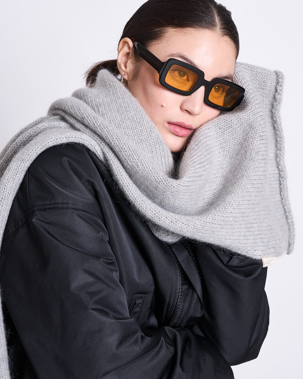 A woman with brown hair wears orange-tinted sunglasses, a black jacket, and the jan-n-june WOOLY KNIT SCARF AVAK LIGHT GREY in a soft wool-mohair blend wrapped around her neck and head, posing against a plain light background.