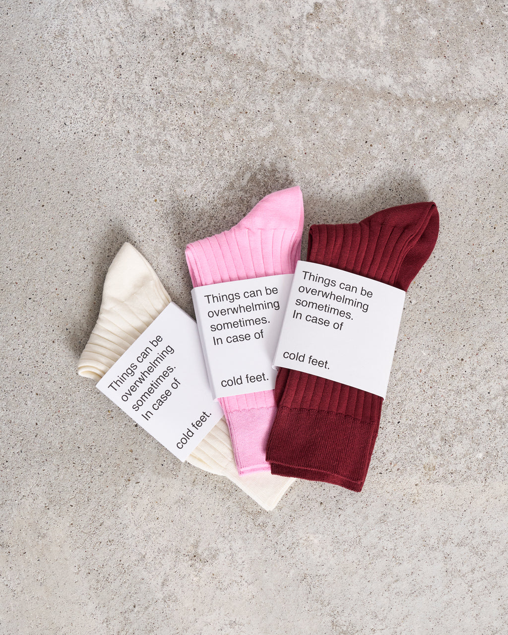 Three pairs of jan-n-june SOCKS OFFWHITE—cream, pink, and burgundy—are displayed on concrete, each wrapped with a label reading “Things can be overwhelming sometimes. In case of cold feet.” Made from soft organic cotton.