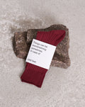A pair of red organic cotton SOCKS SYRAH by jan-n-june, wrapped in a white label with the text: Things can be overwhelming sometimes. In case of cold feet. Cosy colors for those seeking comfy and soft comfort, placed on a stone surface.
