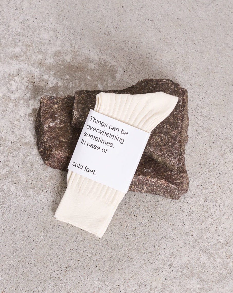 A pair of comfy jan-n-june SOCKS OFFWHITE, made from organic cotton with a label saying Things can be overwhelming sometimes, are shown placed on a rough brown stone atop a concrete surface.
