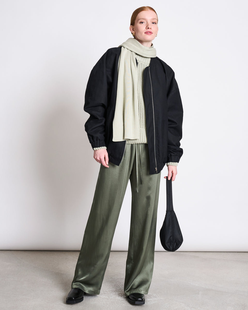 A woman stands against a plain wall wearing sage green satin pants, a cream organic cotton sweater, black jacket and shoes, holding a black bag. She has her hair tied back and wears the jan-n-june KNIT SCARF STATEN VINTAGE LACE GOTS.