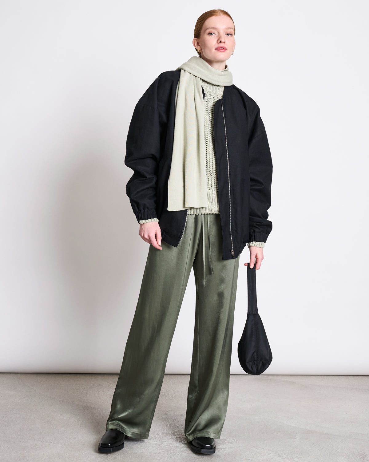A woman stands against a plain wall wearing sage green satin pants, a cream organic cotton sweater, black jacket and shoes, holding a black bag. She has her hair tied back and wears the jan-n-june KNIT SCARF STATEN VINTAGE LACE GOTS.