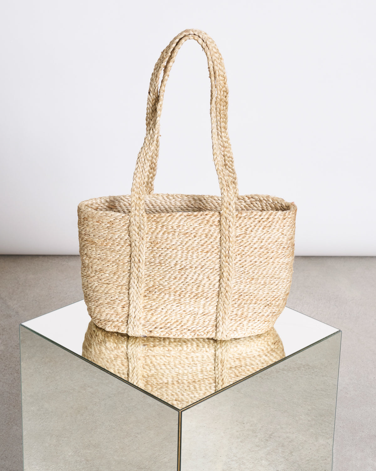 The jan-n-june SHOPPING BASKET JUTE LONG HANDLE, an organic tote with two handles, is displayed on a mirrored cube against a white background, highlighting its craftsmanship and empowerment program origins.