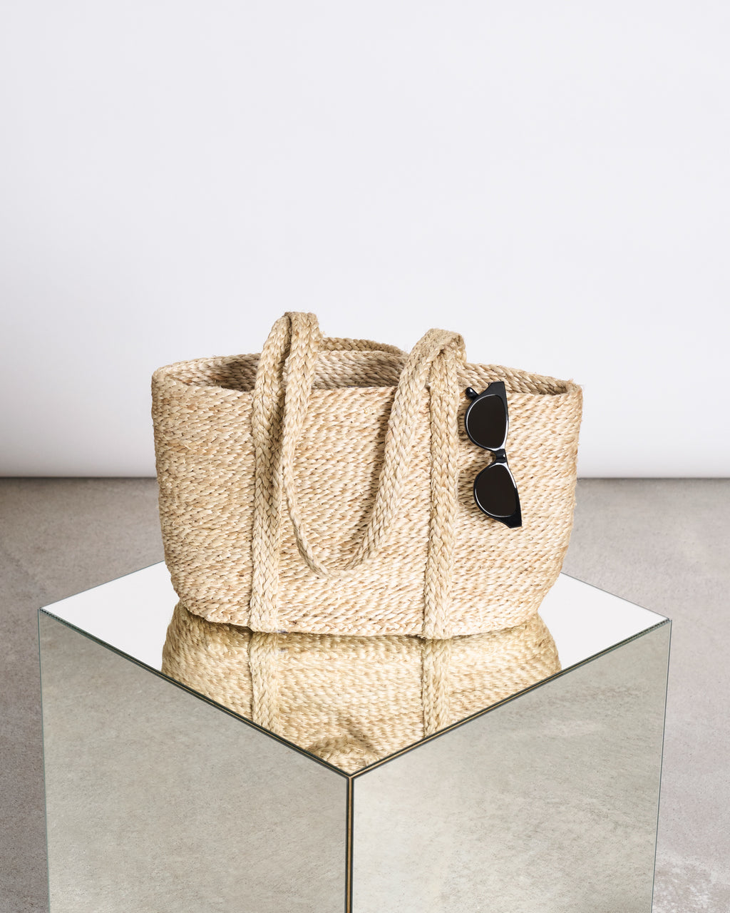 The jan-n-june SHOPPING BASKET JUTE LONG HANDLE rests on a mirrored pedestal, featuring a pair of black cat-eye sunglasses hanging from one handle, set against a minimalistic background.
