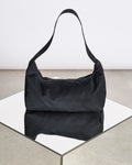 The BAG TUFAO BLACK by jan-n-june, an over-the-shoulder bag with a single strap, is shown on a mirrored surface with its reflection visible. A neutral, minimalistic background highlights its stylish and practical design.