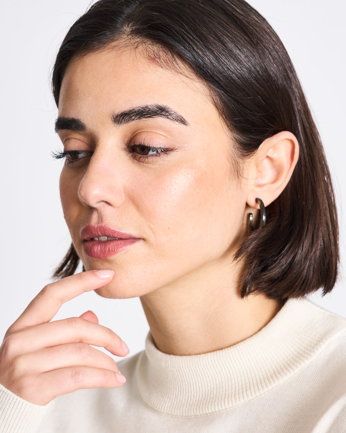 A woman with short dark hair wears jan-n-junes MINI HOOPS MIDNIGHT HORN, gazing downward thoughtfully. Her cream-colored top and calm pose highlight the timeless appeal of her accessory against a plain background.