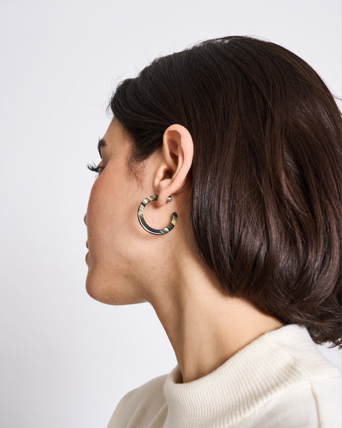 A woman with short dark hair, wearing a cream sweater and jan-n-junes MINI HOOPS MIDNIGHT HORN earrings, is shown in profile against a plain light background.