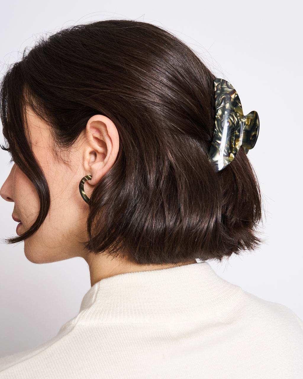 A woman with short brown bob hair, wearing a cream top and gold hoops, styles her hair at the back with jan-n-june’s sustainable MIDI HEIRLOOM CLAW MIDNIGHT HORN, made in Italy and featuring a striking dark green patterned design.