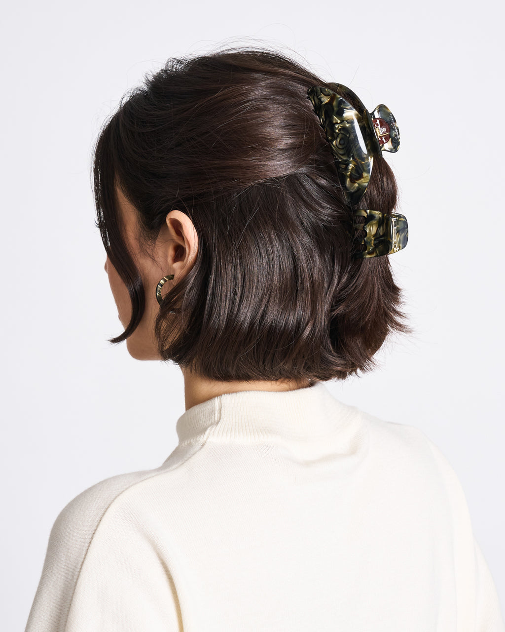 A woman with short, dark brown hair in loose waves wears the jan-n-june MINI CLAW MIDNIGHT HORN, handcrafted in Italy, and a white turtleneck sweater, facing away from the camera against a plain background.
