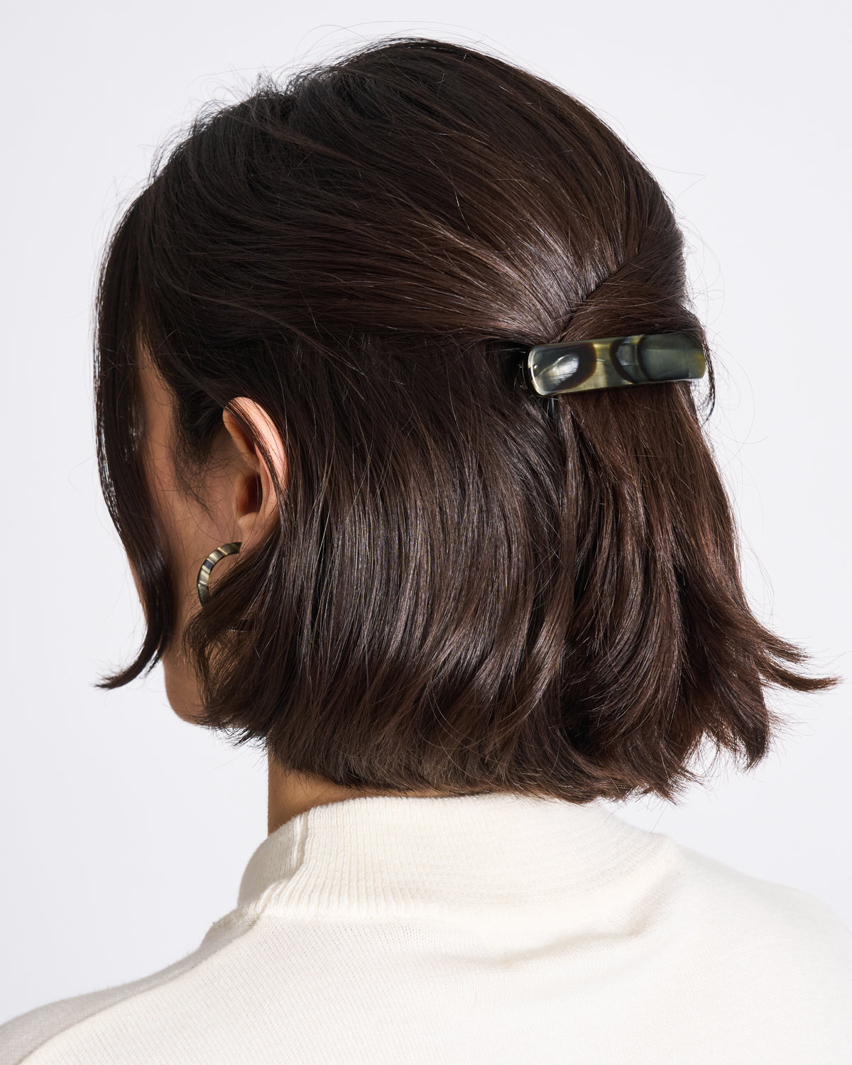 A person with short, dark brown hair, partially pinned back with the jan-n-june HEIRLOOM BARRETTE MIDNIGHT HORN, wears a cream top. The image is taken from behind, emphasizing their French chic hairstyle and the stylish accessory.