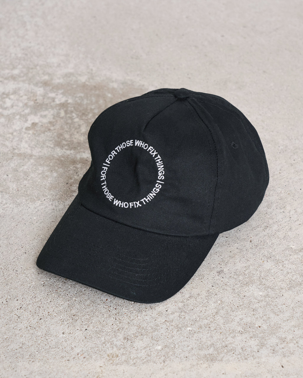 A black organic cotton cap from jan-n-june, the CAP FOR THOSE WHO FIX THINGS BLACK GOTS, rests on concrete with white circular embroidery that reads FOR THOSE WHO FIX THINGS.