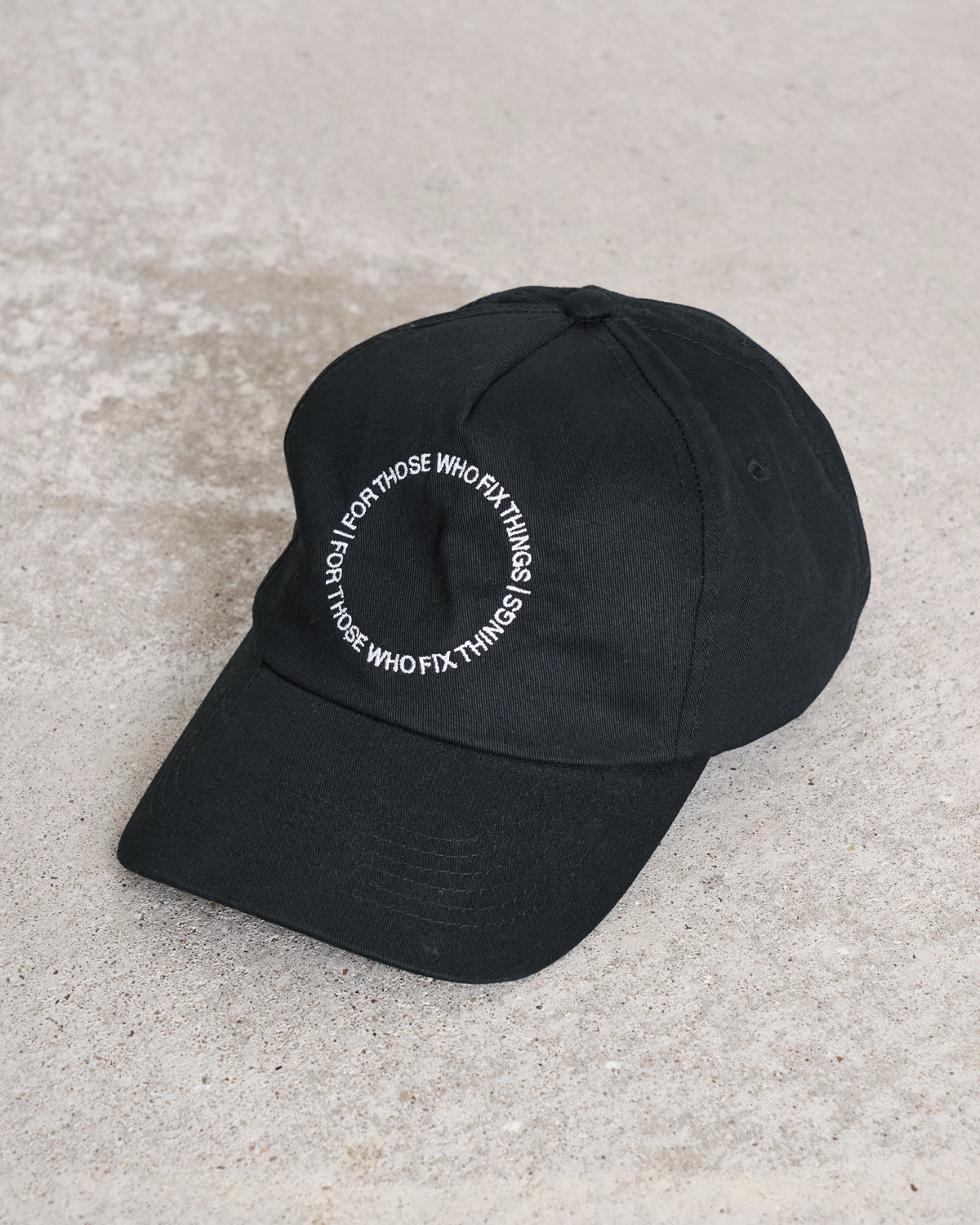 A black organic cotton cap from jan-n-june, the CAP FOR THOSE WHO FIX THINGS BLACK GOTS, rests on concrete with white circular embroidery that reads FOR THOSE WHO FIX THINGS.