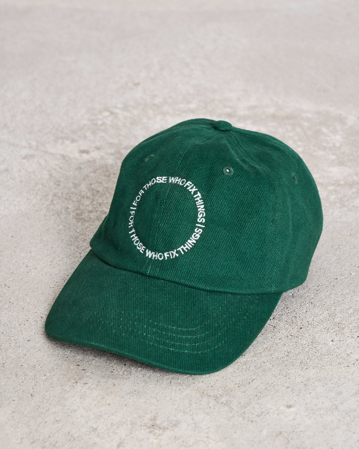 The jan-n-june CAP FOR THOSE WHO FIX THINGS BOTTLE GREEN GOTS is an organic cotton, GOTS certified green cap with white circular embroidered text, displayed on a light gray concrete surface.