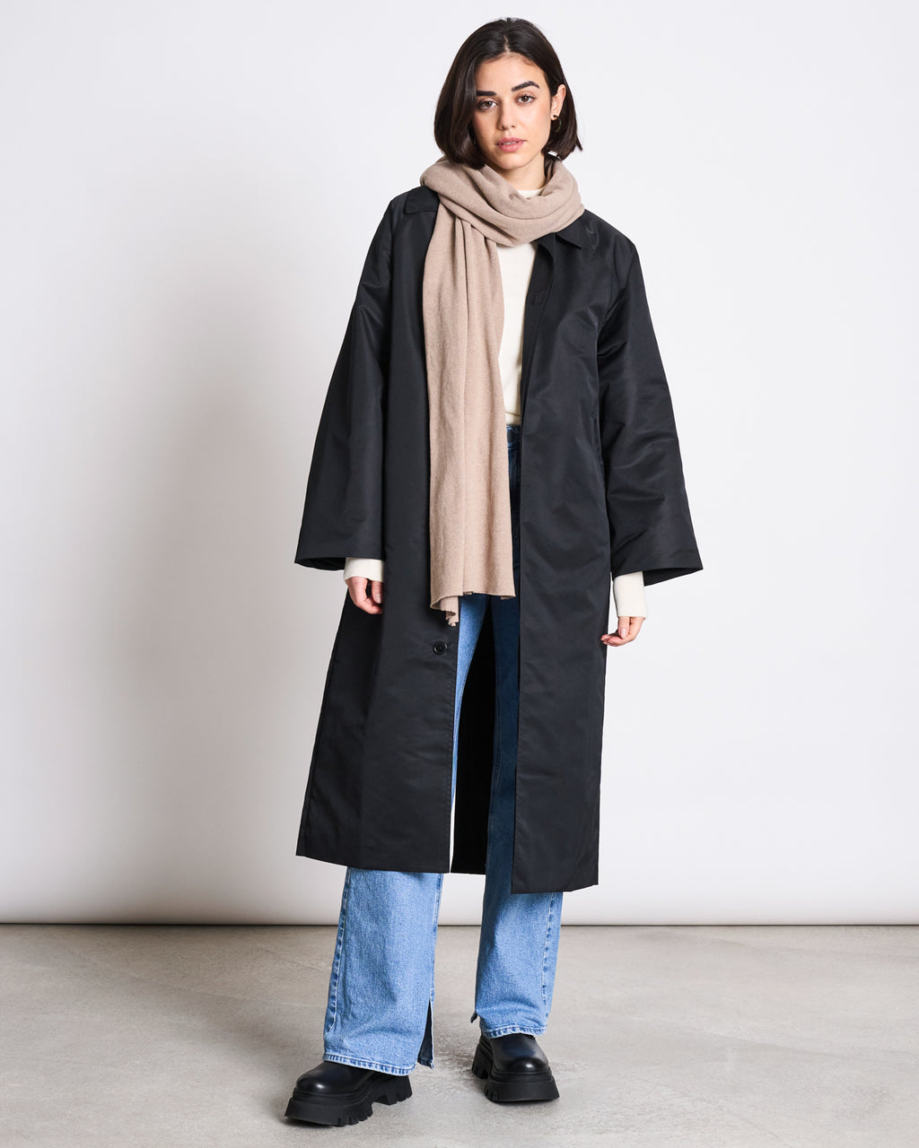 A woman stands against a plain background wearing jan-n-junes SCARF ROSS SAND, a long black coat, white top, light blue jeans, and black platform shoes, with her hands by her sides and a neutral expression.